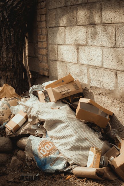 A pile of discarded cardboard boxes, some flattened and others partially intact, resting against a rough, weathered brick wall outdoors. The boxes vary in size, with visible printed labels and brown kraft finishes. Several are partially covered by a large, textured, off-white fabric sack or bag that appears used for waste containment. Surrounding the pile are small stones and dirt, with a nearby tree trunk partly visible on the left edge of the image. The scene is illuminated by natural daylight, casting soft shadows on the objects and the wall, highlighting the textures of the cardboard and fabric. The backdrop consists of a brick wall with sectioned concrete or cinder blocks, suggesting an external environment where private or third-party waste collection might occur instead of municipal disposal. This setting exemplifies areas where independent rubbish collection or on-site clearance services, such as those provided by companies like rubbishclearancefinsburypark.com, could handle waste removal efficiently, particularly in situations involving bulk cardboard and packaging materials in a residential or commercial outdoor area.
