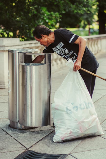 A middle-aged man with dark hair wearing a black T-shirt with white print is seen emptying a large white plastic bag into a metal litter bin on a paved outdoor area. The bin is cylindrical, made of brushed stainless steel with a slot for rubbish, and is positioned next to a low stone or concrete wall with rounded edges. The surrounding environment includes lush green trees and foliage, suggesting a park or public space setting, with natural light illuminating the scene. The man holds a wooden stick or broom handle in his right hand, which is resting against his side, while his left hand guides the bag into the bin. The overall scene reflects on-site or private waste handling, exemplifying a waste clearance activity suitable for independent rubbish collection services. The setting emphasizes the practical aspect of rubbish disposal outside of local authority collection, aligning with professional waste management practices such as those provided by rubbishclearancefinsburypark.com in the context of alternative waste handling options.