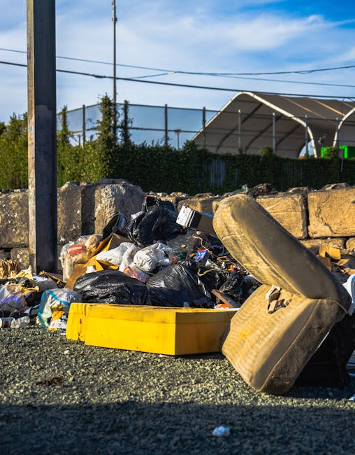 A waste collection worker wearing a high-visibility yellow and red uniform is seen using a small tool to assist in the emptying of a large red rubbish truck parked on the side of a paved road. The truck's rear hatch is open, revealing an internal mechanism for compacting waste. The worker is standing on a paved area next to the truck, with a black garbage bag placed on a small platform at the back. Nearby, a black motorcycle with a metal rack at the rear is parked close to the truck, with some scattered litter nearby. In the background, there are trees, utility poles with power lines, and distant commercial buildings, indicating an urban or suburban environment. The scene is captured during daylight with overcast skies, providing neutral lighting that highlights the textures of the truck's painted metal surface, the worker's safety clothing, and the surrounding street elements. This image relates to private or industrial waste collection services, reflecting the typical activities of rubbish removal operations in the area.