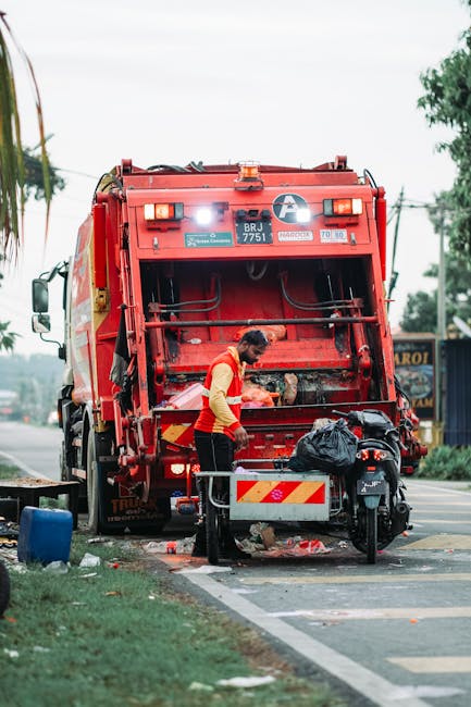 A street scene showing a worker operating a red rubbish collection vehicle parked on the side of the road, with the rear compartment open and partially filled with waste materials. The worker, dressed in a high-visibility yellow and red uniform, is leaning forward, handling an item or cleaning the compartment. Nearby, there is a black rubbish bag resting on a small metal platform attached to the vehicle, and some litter on the pavement, including a black bag and scattered debris. The background includes a tree-lined street with utility poles, a blue building, and traffic on the road, under a cloudy sky. The scene reflects a routine waste collection operation, typical of private rubbish removal services, with emphasis on efficient on-site clearance and waste management support in an urban environment.