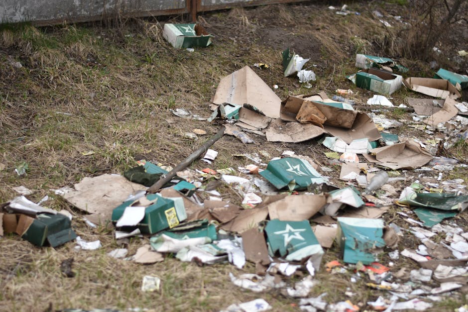 An outdoor scene showing scattered cardboard pieces, some torn and crumpled, along with broken paper packaging and plastic debris on the ground near a grassy embankment. The cardboard includes remnants of green and white printed boxes, possibly from beverage or food packaging, with visible logos and branding. The debris appears to be mixed with other litter such as small paper fragments and plastic bottles. The setting appears to be a neglected or unmanaged area, with no visible waste collection containers nearby. The uneven ground features patches of grass and dirt, with a slight incline leading up to a wooden fence at the back. The overall environment suggests a site where private rubbish has been improperly discarded, highlighting the importance of professional rubbish removal services like those offered by [COMPANY_NAME] for effective waste management and site clearance.