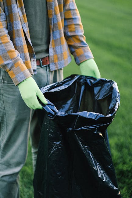 A person wearing a yellow and blue checkered flannel shirt, grey trousers, and bright green gloves is holding open a large black plastic rubbish bag outdoors on a grassy area. The bag appears to be ready for waste collection and is positioned in front of the person's waist, with their hands grasping the top edge. In the background, there is a lush green lawn, suggesting a garden or park setting, which relates to waste removal or rubbish clearance activities. The scene emphasizes private waste handling, with the individual preparing the bag for rubbish disposal, potentially as part of an independent collection or on-site clearance process, aligning with services such as those offered by rubbishclearancefinsburypark.com.