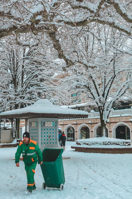 A waste collection worker dressed in a green and orange uniform is walking through a snowy outdoor area, pushing a green wheeled bin on a paved surface covered with snow. The background features a small pavilion with a tiled roof, supported by white columns, and a series of brick and plaster buildings with arched entrances. Tall, leafless trees with snow-covered branches surround the scene, creating a wintery atmosphere. The worker appears to be engaged in rubbish collection as part of an independent waste removal service, reflecting a winter environment where private waste handling continues despite snowy conditions. The scene is well-lit by natural daylight, emphasizing the crisp white snow and the contrasting dark branches of the trees. The setting suggests an urban or suburban public space, possibly a park or communal area, where rubbish removal activities are being carried out in winter weather.