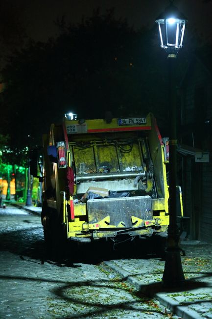 A small yellow and black rubbish collection vehicle is parked on a cobbled street at night, adjacent to a traditional black street lamp illuminating the scene. The vehicle's rear panel is open, revealing collection equipment and some waste materials. The vehicle appears to be used for private waste clearance, situated on a narrow residential or urban street with cobblestone paving. In the background, faintly illuminated by nearby streetlights, there are trees and possibly building outlines, contributing to a quiet, nocturnal atmosphere. The scene reflects a setting for independent waste removal services, such as those provided by companies like rubbishclearancefinsburypark.com, operating outside of official council refuse collection schedules, with the urban environment enhanced by soft and directional lighting and the textured surface of the cobbles.
