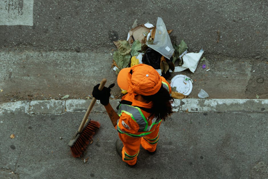 A sanitation worker dressed in an orange high-visibility uniform with reflective stripes, wearing a matching orange cap and black gloves, is standing on a concrete pavement beside a street curb. The worker is holding a large broom with a wooden handle and red bristles, positioned upright with the bristles touching the ground. In front of the worker, there is a pile of mixed rubbish on the edge of a concrete wall, which includes crumpled paper, plastic packaging, a white disposable plate, and leafy plant debris. The scene is outdoors, with natural light illuminating the area, and the background features a weathered concrete wall that borders the street. This image relates to private waste clearance services, showing a waste management operative engaged in rubbish collection as part of an independent disposal or clearance process often seen in urban environments.